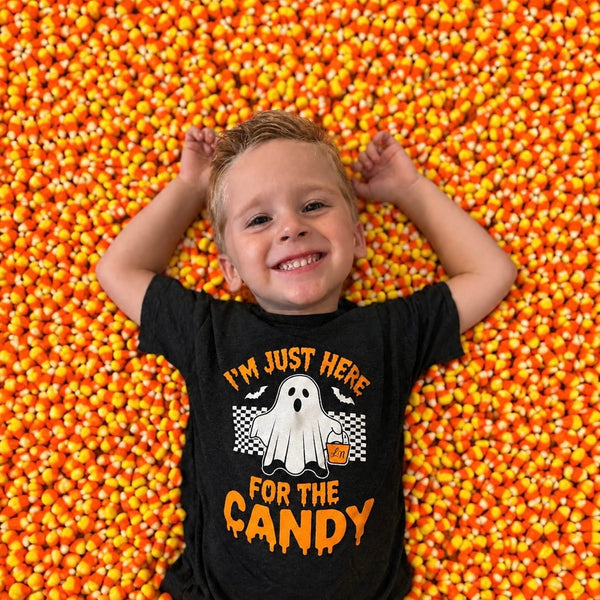 A smiling young boy lies on his back in a ball pit filled with orange and yellow candy corn, wearing a black graphic t-shirt that reads 'I'M JUST HERE FOR THE CANDY' with a ghost illustration.