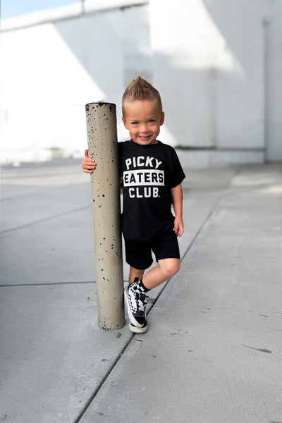 Child wearing a black t-shirt with 'Picky Eaters Club' text, standing next to a concrete pole.