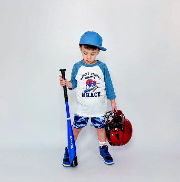 Child holding a baseball bat and helmet, wearing a baseball-themed shirt that says WIggity Wiggity Whack 